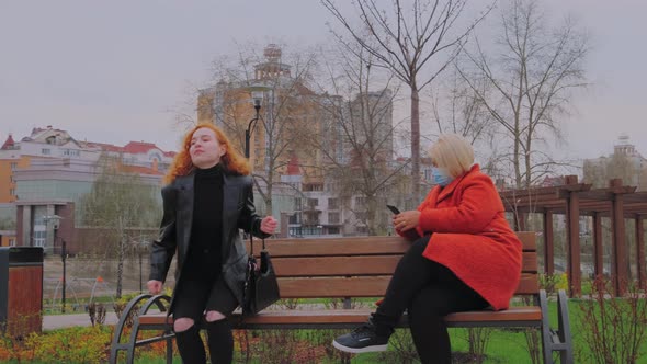 Woman Without Protective Mask Sits Down to Woman in Mask in the Park alt