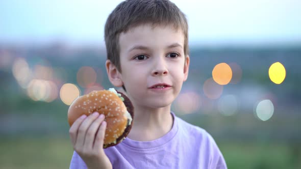 A Child Eats a Big Burger in Nature Against the Background of a Defocused City