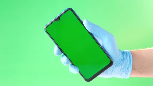 Hand in Latex Gloves Holding Smart Phone Against Green Background alt