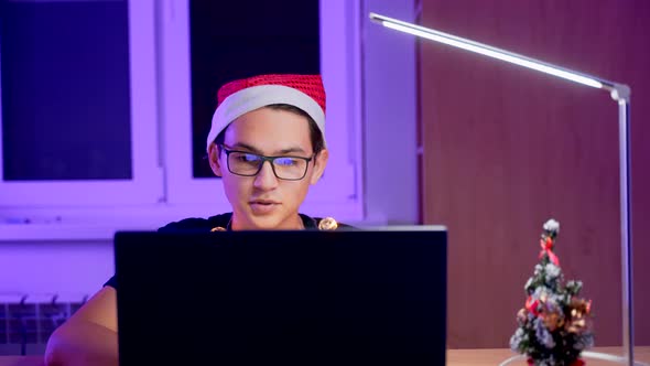 A Young Man in Glasses and a New Year's Hat with a Laptop at the Table. A Student on New Year's Eve alt