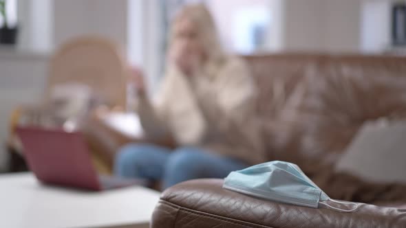 Closeup Coronavirus Face Mask Lying on Couch with Blurred Woman Entering Sitting Down Working Online alt