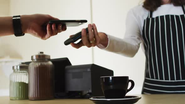Midsection of caucasian customer, using smartphone to pay bill to waitress holding payment terminal alt