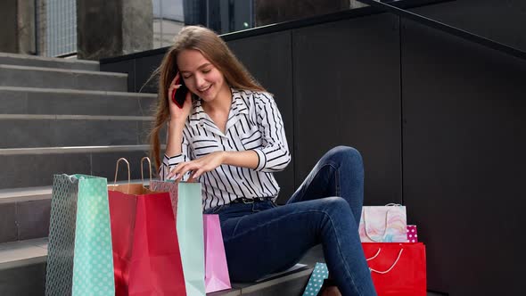 Girl Sitting on Stairs with Bags Talking on Mobile Phone About Sale in Shopping Mall in Black Friday alt