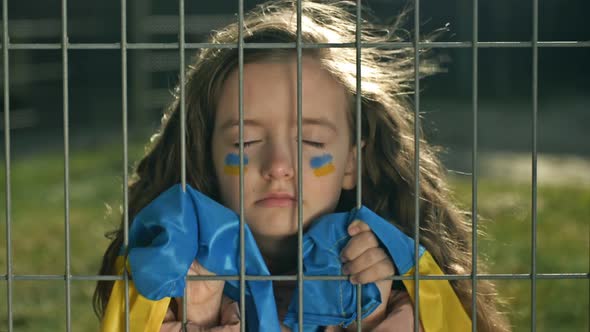 Little Girl is Covered with a Ukrainian Flag with Her Eyes Closed Behind a Metal Grill alt