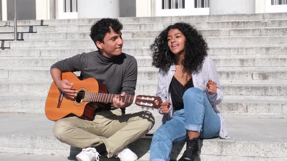 A young latin couple playing the guitar and singing sitting on stairs. alt