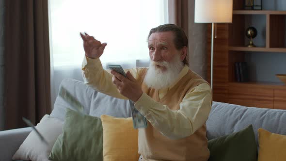 Excited Senior Bearded Man Throwing Dollar Banknotes in Air Sitting on Sofa at Home Enjoying Win or alt