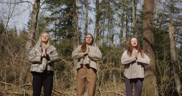 three girls meditate in the forest, inhale the air and slowly exhale raising their hands up alt