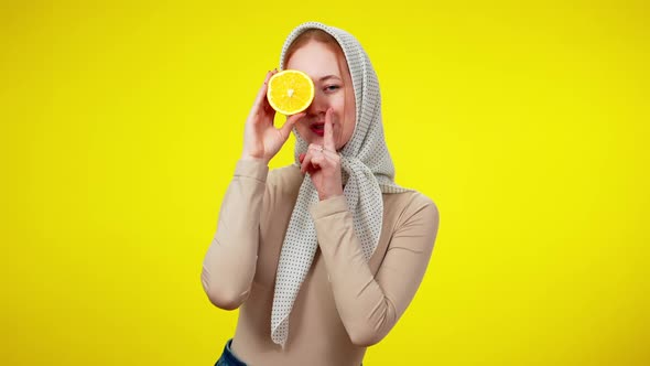 Cheerful Redhead Woman Closing Eye with Half of Orange Putting Finger on Lips alt
