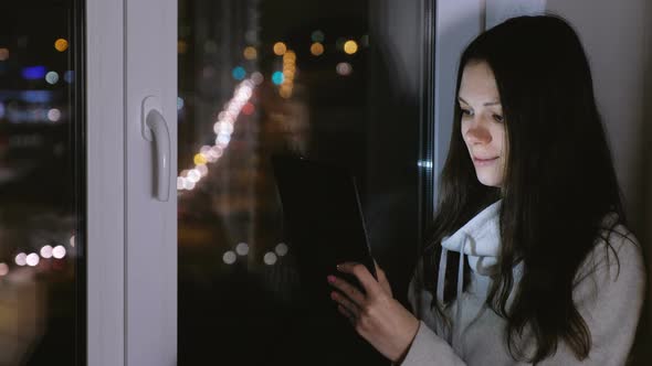 Woman Is Watching Film on Tablet Computer and Laughing Sitting on the Windowsill in the Dark Night
