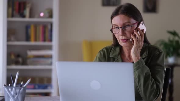 Beautiful Mature Businesswoman is Talking on Phone and Working with Laptop at Table at Home Spbi alt