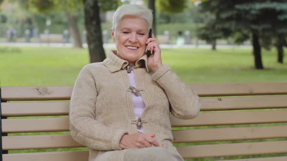 Happy Elderly Woman with Beautiful Face Sitting on Park Bench Takes Phone Call Grayhaired Lady of alt