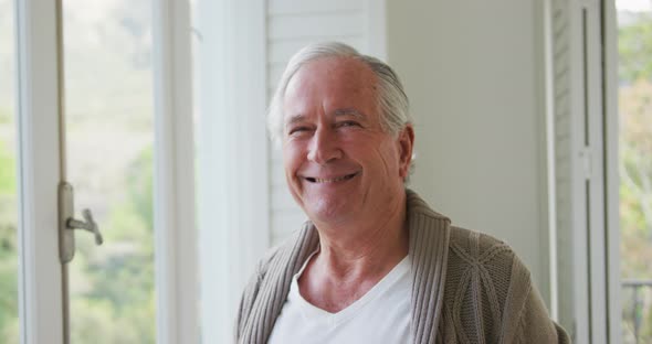 Portrait of cheerful senior man in cardigan sweater standing by window at home alt