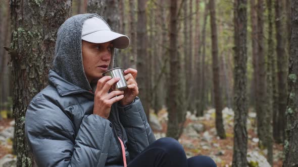 Portrait of Young Woman Hiker Drinking Tea From Metal Cup Sitting in Forest