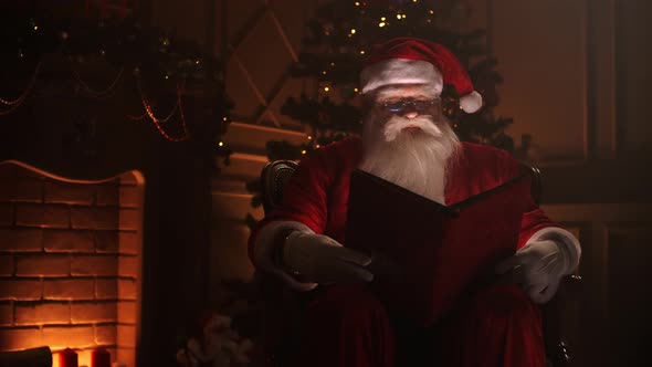 Joyful Santa Clause Sitting in His Rocker in Decorated Room, Reading a Book with Red Cover - Holiday alt