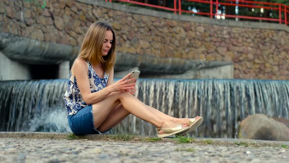 Girl Sitting On Cobblestone Quay In City Park Near Waterfall And Reading News On Mobile Phone