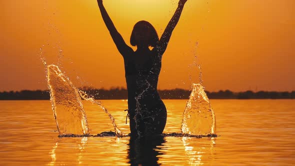 Silhouette of Woman at Sunset Raises Hands Up and Creating Splashes of Water. Slow Motion alt