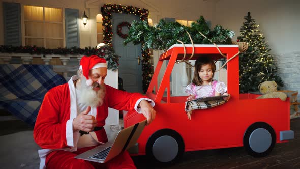 Santa Claus Makes Thumbs Up Working with Laptop and Little Girl Enjoying with Presented Tablet.
