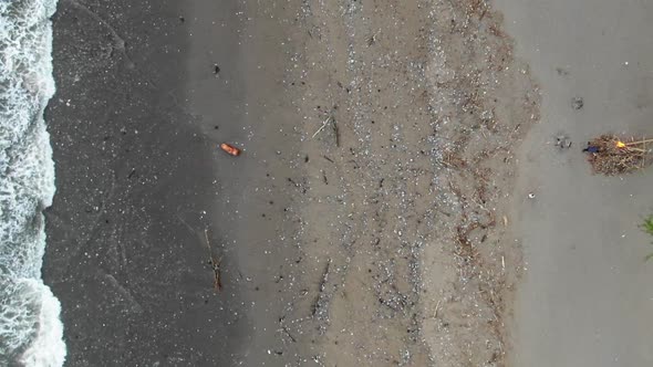 Descending aerial shot of a polluted beach, as people passing by, waves washing out plastic garbage alt