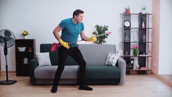 A Caucasian man plays a virtual guitar with a mop, dances and sings and imagines a concert