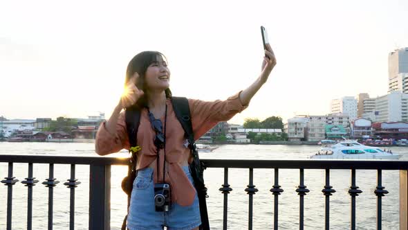 Asian woman tourist with backpack taking selfie and waving on river wharf in Bangkok, Thailand