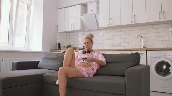 Young Pretty Woman with Pink Hair Watching TV on Sofa in Kitchen Switch Channels alt