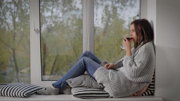 Young Woman Sitting at Windowsill with Glass of Red Wine alt