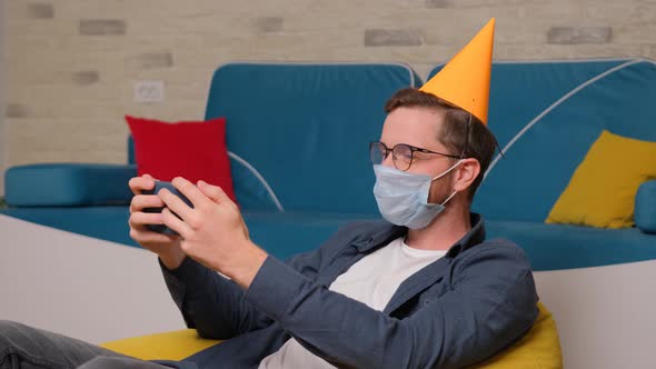 A Young Man with a Birthday Hat and a Medical Mask on His Face Talks Online with His Friends. alt