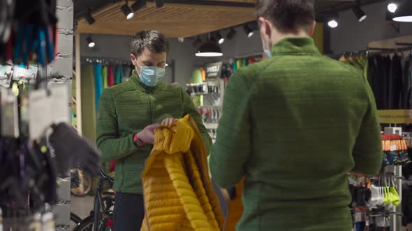 Masked Man in Sports and Outdoor Equipment Store Selects Jacket During Covid 19 Quarantine and Keeps alt