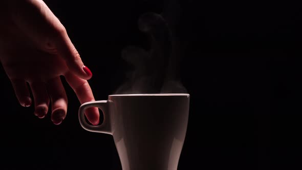 Close-up female's hand takes a white cup with hot coffee or tea on black background with light.