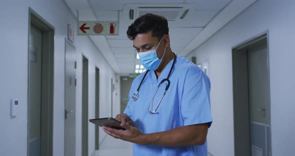 Mixed race male doctor wearing face mask standing in hospital corridor using tablet alt