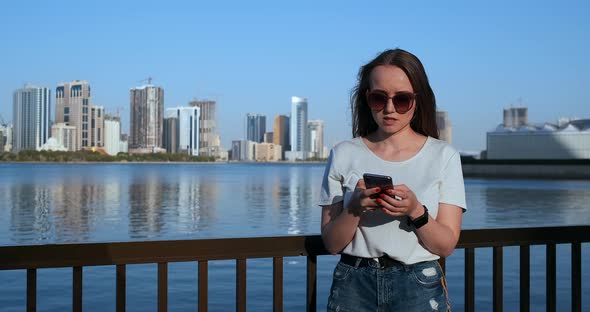 Beautiful Girl with Long Hair in Sunglasses Using Smartphone App at Sunset River Quay Near alt