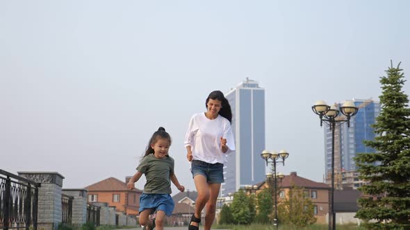 Smiling Asian Girl with Mother Runs Along City Embankment alt