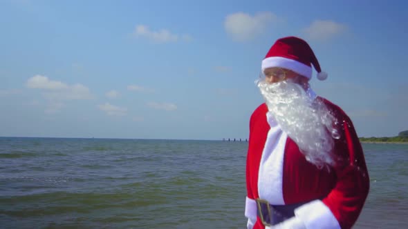Santa Claus in costume walks along sandy shore of sea alt