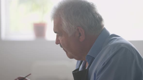 An Elderly Man in an Apron Paints a Picture with a Brush While Sitting at the Table alt