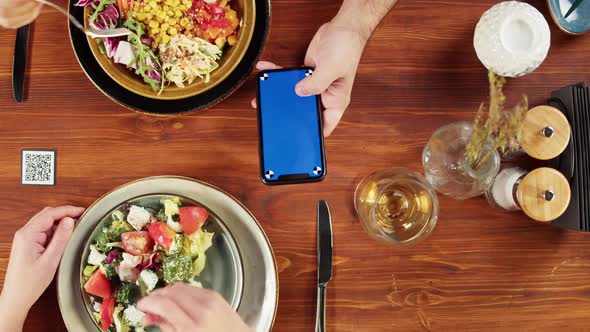 Woman Using Smartphone with Blue Screen and Eating Poke Bowl Salad Qr Code with Online Menu in Cafe alt