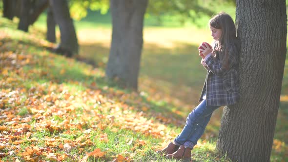 Girl in a Coat stands Leaning on a Tree in a Sunny Autumn Park and Eats a Ripe Apple