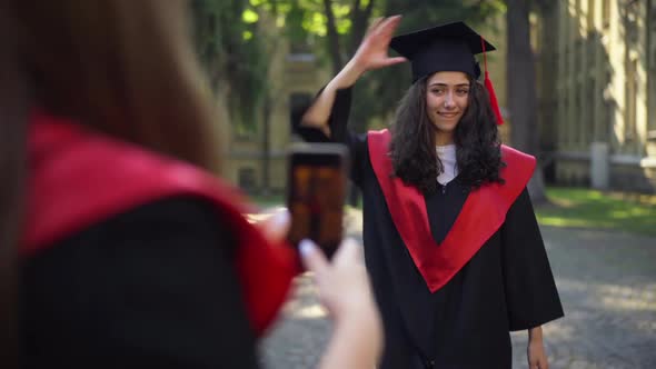Cheerful Graduate Woman Throwing Mortarboard Cap As Friend Taking Photo on Smartphone alt