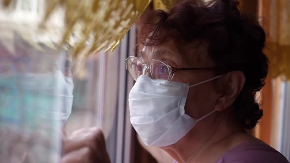 Home Isolated. An Elderly Woman in a Protective Medical Mask Looks Out the Window with Sadness