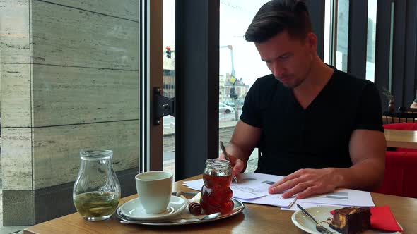 A Young Handsome Man Sits at a Table with Meal in a Cafe and Does Paperwork alt
