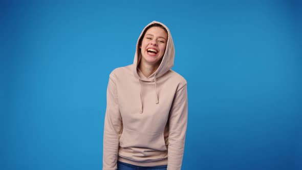 Young Happy Woman in Beige Hoodie Laughing Against Blue Background