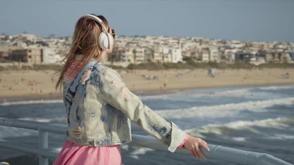Back View of Joyful Woman with Blowing Hair Stylish Clothes on Beach Pier RED alt