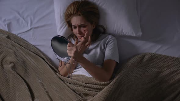 Crying Female Covering Face Wound With Foundation Looking in Mirror Lying in Bed alt