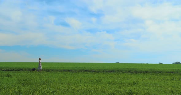 A Girl in a Dress and a Hat Walks Through a Green Field of Grass