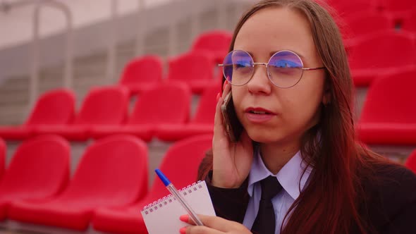 Young Woman in Glasses with Notepad Pen Talking on Mobile Phone Sitting on Stadium Bleachers alt