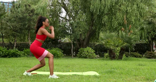 Fitness trainer with a rubber sports belt on hips performs lunges while standing on green grass.