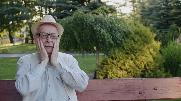 Senior Man Corrects Glasses and Rubs His Head and Face From Fatigue on Bench alt