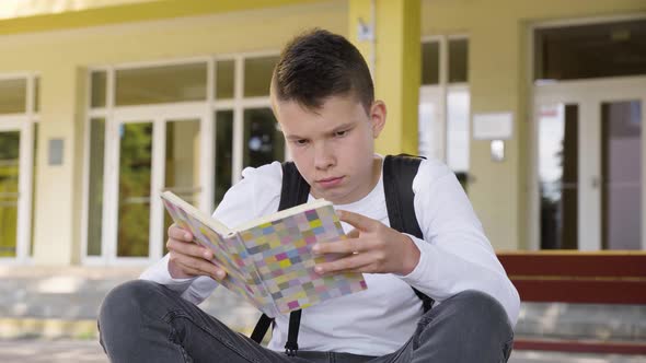 A Caucasian Teenage Boy Reads a Book As He Sits in Front of School  Closeup