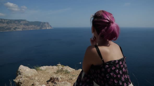 A Girl with Purple Hair Sits on a Hillside Against the Backdrop of a Seascape
