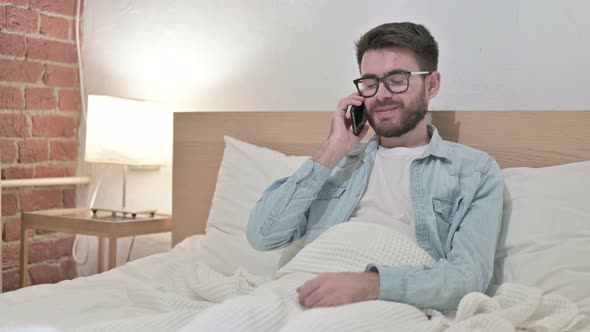Cheerful Young Designer Talking on Smartphone in Bed alt