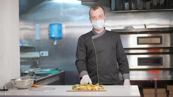 Confident Positive Caucasian Man in Coronavirus Face Mask Posing with Cooked Pizza in Kitchen alt